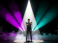 A singer gripping a microphone stand at center stage, bathed in a cone of white spotlight against a dark concert backdrop. Colored stage lights in purple and teal fan outward from behind, creating a dramatic halo effect. Smoke drifts low across the stage floor. Style: concert photography, photorealistic. Mood: dramatic and electrifying.