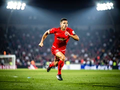 An athlete in a bright red jersey sprinting down a grass football field, arms pumping, stadium crowd blurred in the background under bright floodlights. Sharp directional overhead lighting highlights the motion and muscle definition while the background fades into a soft bokeh. Style: high-speed sports photography. Mood: intense and dynamic.