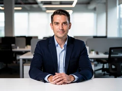 A confident professional man in a navy blazer seated at a clean modern desk, looking directly at the camera with a composed expression, a blurred open-plan office visible behind him. Soft diffused studio lighting from the front illuminates his face evenly with no harsh shadows. Style: corporate portrait photography. Mood: polished and approachable.