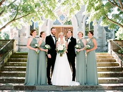 A wedding party of six people standing in a row on stone steps outside a sunlit chapel, dressed in formal attire — the bride in white, groom in black, and four attendants in matching sage green. Bright midday sunlight filters through surrounding trees, creating gentle dappled highlights. Style: classic wedding photography. Mood: joyful and celebratory.