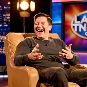 A person relaxed in a tan leather guest chair on a late-night talk show set, laughing mid-conversation with one hand gesturing expressively. Warm studio spotlights illuminate their face from above and slightly to the left, creating a flattering soft shadow under the chin. A colorful backdrop with the show's logo glows behind them. Style: broadcast television photography. Mood: charming and spontaneous.