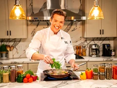 A cheerful person in a white chef's apron standing behind a marble kitchen counter, tossing fresh herbs into a sizzling pan with a confident grin. Warm overhead pendant lights cast a golden glow on the food and their face. Colorful vegetables and spice jars are arranged neatly on the counter. Style: bright lifestyle food television. Mood: warm and inviting.